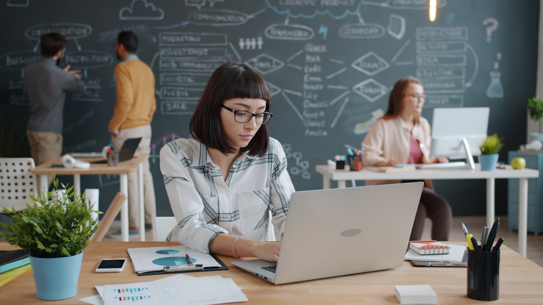 Attractive girl businesswoman is working with laptop typing in coworking place with people multi-ethnic group are visible in background. Youth and business concept.